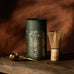 A tub of 'Matcha + Collagen' with a whisk and ritual scoop on a wooden surface